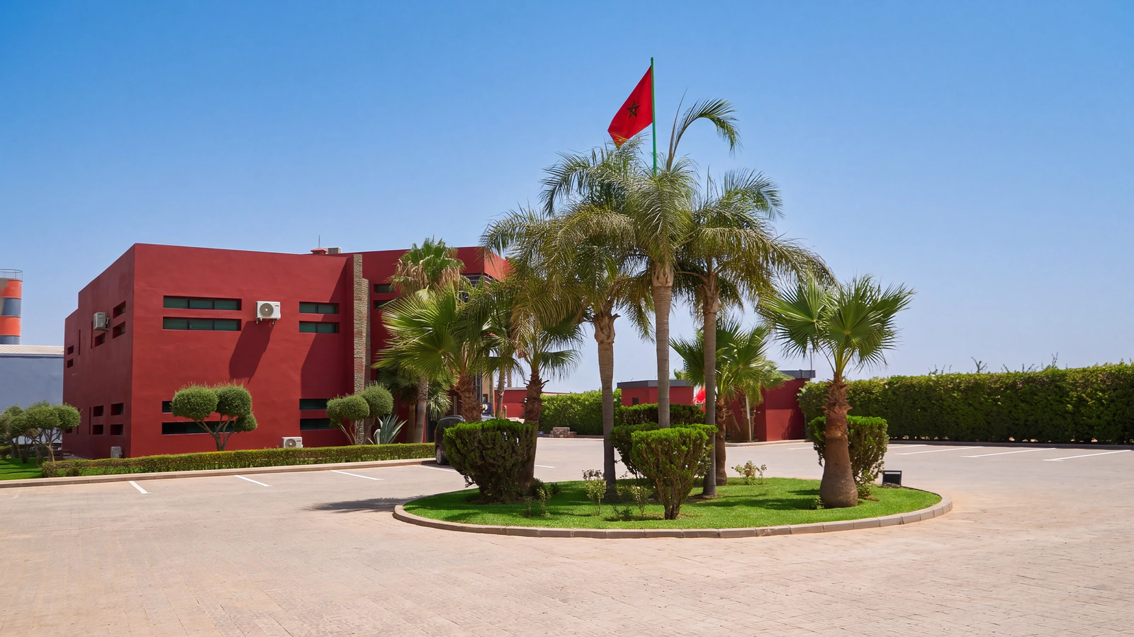 Site de Belza Groupe à Ben Guerir : bâtiment rouge, rond-point planté de palmiers et drapeau marocain