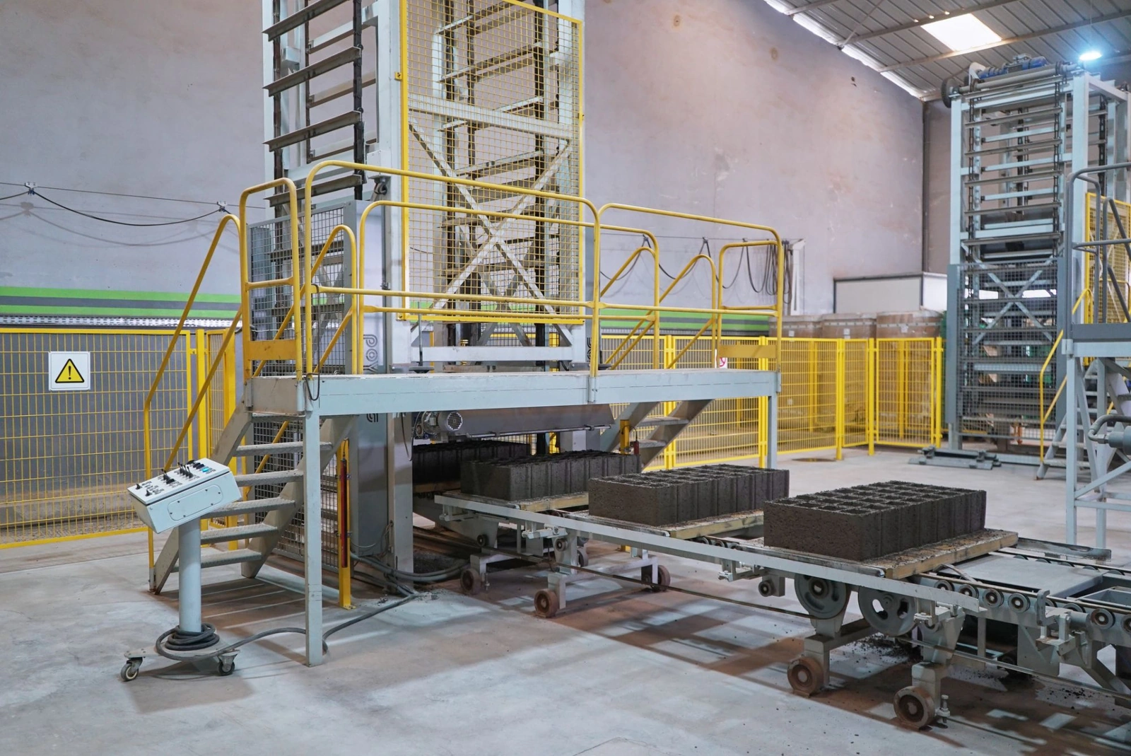 Ligne de production de Belza Ecotech : passerelle jaune, convoyeur de blocs creux frais et empileur